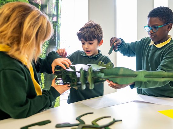Three children in green tops stand around a table building a model minibeast out of flat, green, wooden parts.