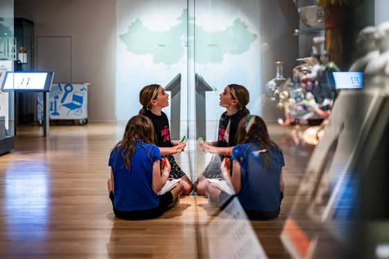 Two children sit on the floor in a museum gallery, looking at objects in a glass display case.