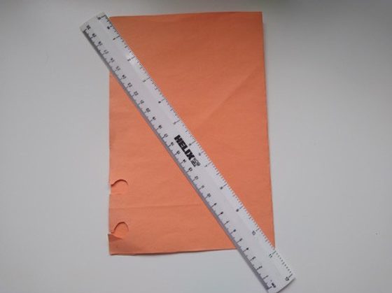 An orange paper folded in half, lying on a flat surface with a transparent long ruler placed across it.