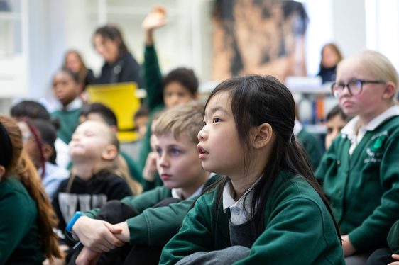 A group of school children sit on a floor listening to a speaker.
