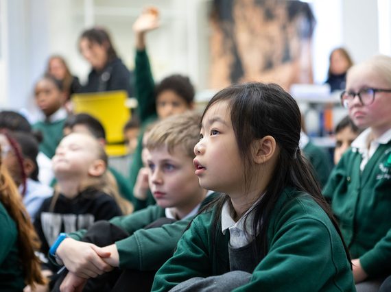A group of school children sit on a floor listening to a speaker.