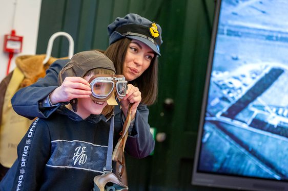 An adult in an RAF costume and hat helps a child try on flight goggles and a flight cap.