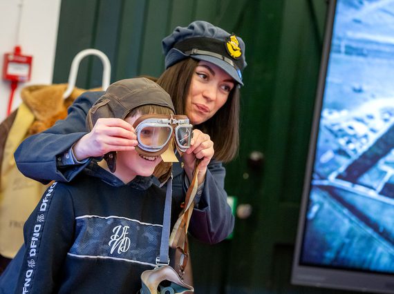 An adult in an RAF costume and hat helps a child try on flight goggles and a flight cap.