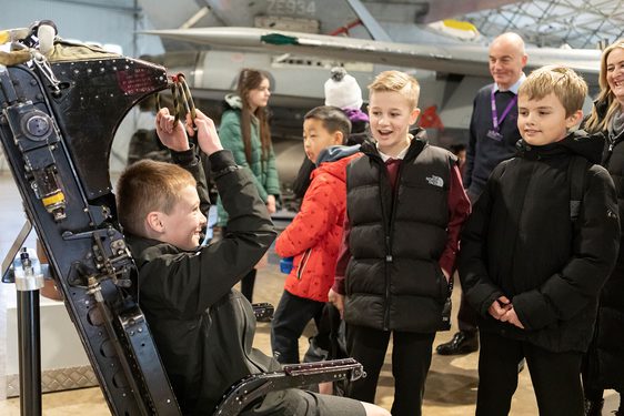 A school pupil sitting in a pilot seat with other pupils looking on.
