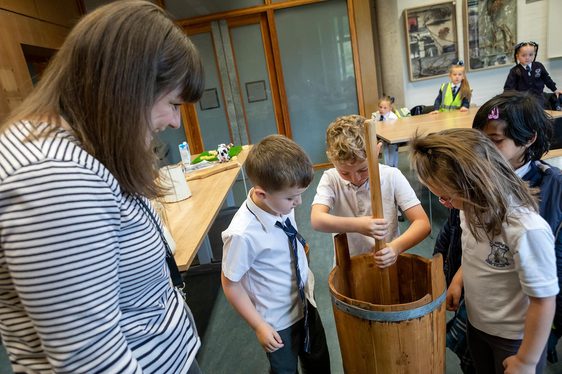 A group of school children churn butter in a classroom with a teacher watching over them.
