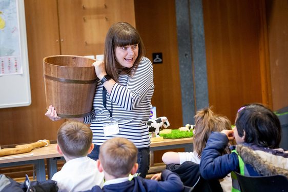 A teacher holding a wooden bucket while talking to a class of children.