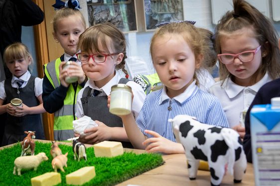 A group of school children hold jars of butter while looking at toy farm animals on a table.