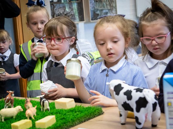 A group of school children hold jars of butter while looking at toy farm animals on a table.
