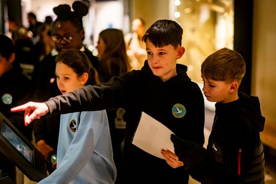 Three school children stand around an interactive screen in a busy gallery. The child in the middle is pointing to something out of frame.