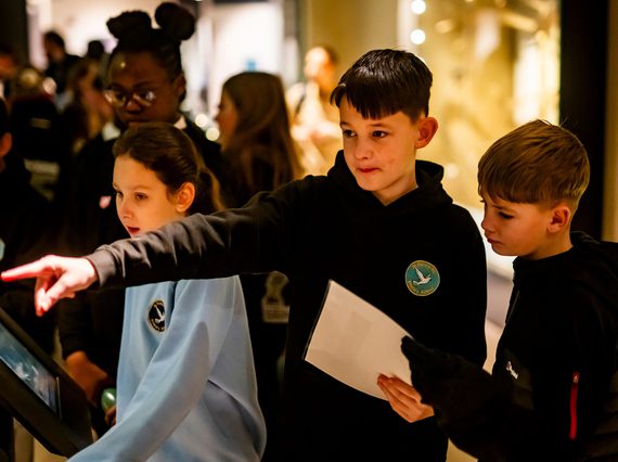 Three school children stand around an interactive screen in a busy gallery. The child in the middle is pointing to something out of frame.