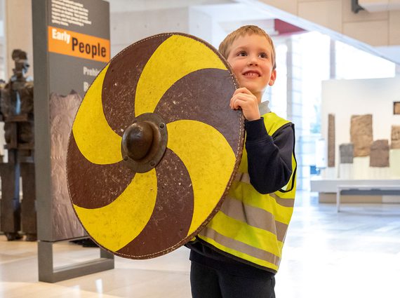 A child in a high-vis vest stands in a gallery space holding a large, round, gold and yellow shield.