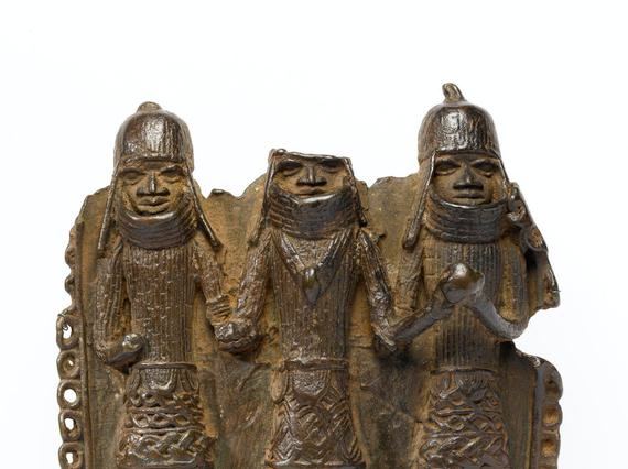 Bronze pendant chipped around the edges. Three figures in armour and heavy helmets stand side by side. The top of the middle figure's head is broken off.